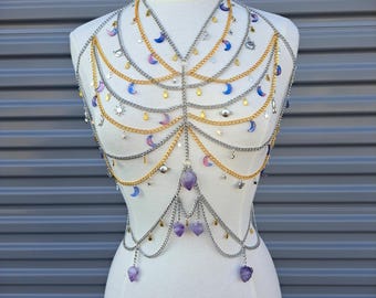 May include: A celestial-themed body chain with silver and gold chains, adorned with crescent moons, stars, and purple gemstone pendants. The layered design drapes over a mannequin, showcasing the intricate details of the jewelry.