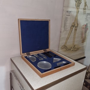 May include: A wooden box with a blue velvet lining, containing a set of clear glass prisms and lenses. The box is open and the prisms are arranged in a grid pattern.