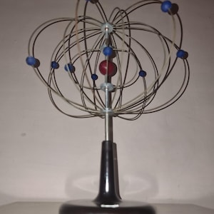 May include: An atom model with a black base and a silver metal rod supporting a wire frame. The wire frame has blue and red spheres representing electrons and the nucleus. This model is a scientific or educational item.