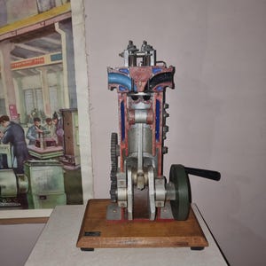 May include: A vertical cross-section model of a combustion engine mounted on a rectangular wooden base. The engine features exposed internal components painted in red, blue, and grey, with a black hand crank attached to the side for manual operation.