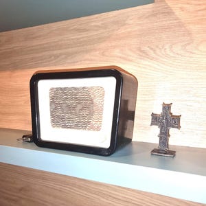Può includere: Una radio vintage nera con una griglia dell'altoparlante bianca si trova su uno scaffale di legno accanto a una piccola croce di bronzo.