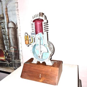 May include: A cutaway model of a single-cylinder internal combustion engine mounted on a rectangular wooden base. The engine features a red cylinder head, light blue internal components, and a silver metal exterior with cooling fins.