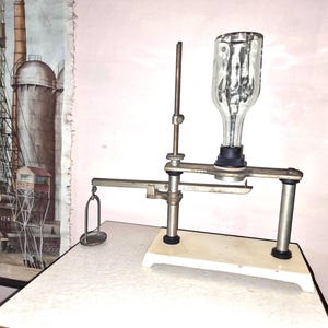 Könnte beinhalten: Eine silberne und gläserne Laborwaage mit einer durchsichtigen Glasflasche auf einem Ständer. Die Waage hat einen Metallarm mit einer Schale an einem Ende und einer Metallstange am anderen Ende. Die Waage steht auf einer weißen Oberfläche.