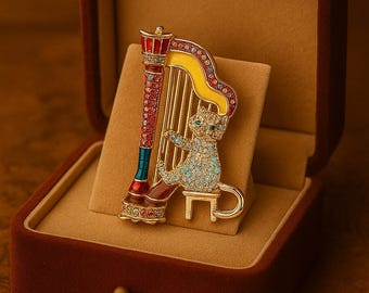 Op de afbeelding: Een met juwelen bezette kat die een harp speelt, broche, gepresenteerd in een fluwelen sieradendoos. De broche heeft een goudkleurige harp met een gele bovenkant en een rode en blauwe kolom. De kat is versierd met blauwe en heldere kristallen.