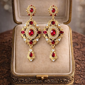 Könnte beinhalten: Ein Paar verzierte, goldfarbene Ohrringe mit roten und klaren Edelsteinen, präsentiert in einer Vintage-Schmuckschatulle. Die Ohrringe haben einen zentralen ovalen roten Stein, der von kleineren klaren Steinen und zusätzlichen roten Akzenten umgeben ist.