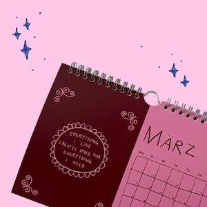 May include: A spiral-bound wall calendar with a pink background and dark blue star illustrations. The left page is deep red with pink decorative swirls and the text: EVERYTHING I LOSE CREATES SPACE FOR EVERYTHING I NEED. The right page is pink with the month M&Auml;RZ.