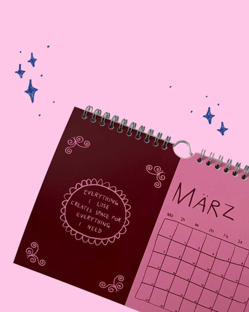 May include: A spiral-bound wall calendar with a pink background and dark blue star illustrations. The left page is deep red with pink decorative swirls and the text: EVERYTHING I LOSE CREATES SPACE FOR EVERYTHING I NEED. The right page is pink with the month M&Auml;RZ.