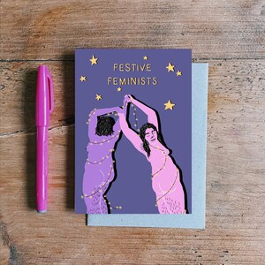 May include: A purple card with two women in pink outlined in black, holding up strings of lights. The text "FESTIVE FEMINISTS" is in gold lettering on the card.