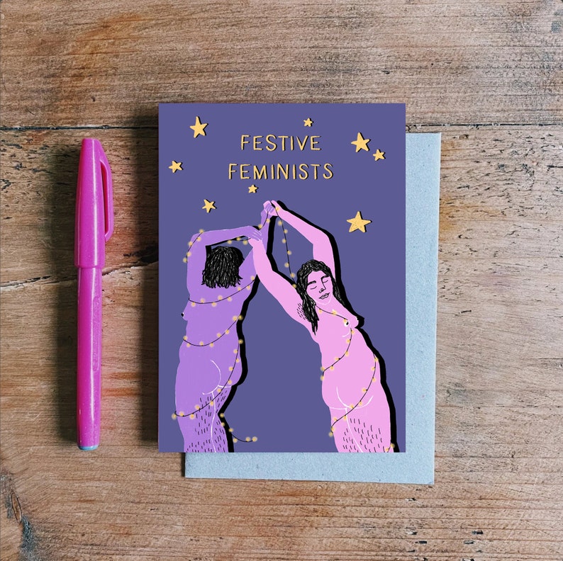 May include: A purple card with two women in pink outlined in black, holding up strings of lights. The text "FESTIVE FEMINISTS" is in gold lettering on the card.