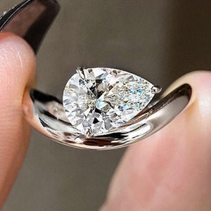 May include: A pear-shaped diamond engagement ring set in a white gold band. The ring is designed with a twisted band and a small diamond accent on the side.