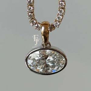 Puede incluir: Un collar de tenis dorado con una hilera continua de pequeñas piedras redondas transparentes. Un gran colgante ovalado con una piedra transparente está engastado en oro en el centro. El colgante tiene un pequeño logotipo con las letras FJ.