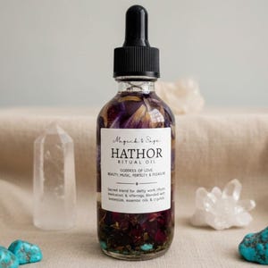 May include: A clear glass dropper bottle filled with dark liquid and dried flower petals. The white label reads Magick & Sage HATHOR RITUAL OIL, GODDESS OF LOVE, BEAUTY, MUSIC, FERTILITY & PLEASURE, and a sacred blend for deity work, offerings, and rituals.
