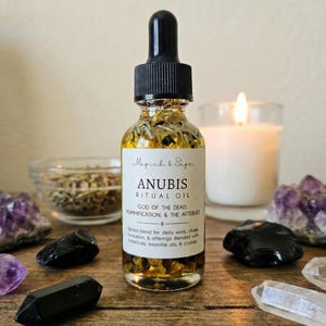 May include: A clear glass dropper bottle filled with golden oil and dried herbs. The white label reads Magick & Sage ANUBIS RITUAL OIL GOD OF THE DEAD, MUMMIFICATION, & THE AFTEBUFE. Sacred blend for deity work, rituals, invocation, & offerings.