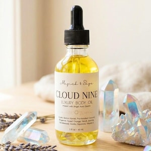 May include: A clear glass bottle of Cloud Nine luxury body oil with a black dropper cap, featuring a label with the text Magick & Sage, Cloud Nine Luxury Body Oil, and 2 fl oz 60 ml. The bottle is surrounded by iridescent quartz crystals and dried lavender sprigs.
