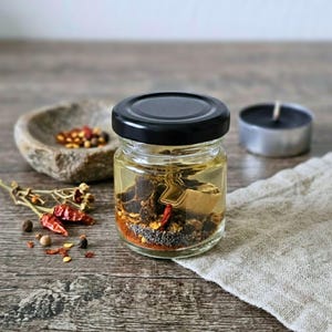 May include: A small clear glass jar with a black lid containing a mixture of oil, dried red chili peppers, dark seeds, and various spices. The jar is positioned on a wooden surface alongside loose dried chili peppers and seeds.