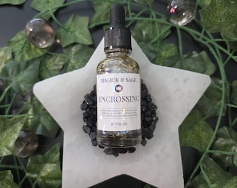 May include: A clear glass dropper bottle filled with liquid and herbs, featuring a white label with the text MAGICK & SAGE, UNCROSSING, and RITUAL OIL. The bottle sits on a pile of small black stones atop a white star-shaped tray.