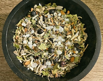 May include: A black circular bowl filled with a dry herbal mixture containing green leaves, small brown twigs, dark purple round berries, and coarse white salt crystals. The contents are a blend of various dried botanical elements and minerals.
