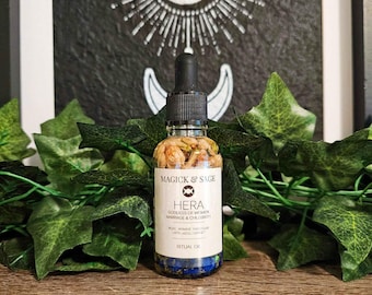 Op de afbeelding: Een doorzichtig glazen druppelflesje gevuld met rituele olie met gedroogde bloemen en blauwe stenen. Het witte etiket bevat de tekst Magick & Sage, Hera, Goddess of Women, Marriage & Childbirth, Rose, Jasmine, Patchouli, Lapis Lazuli, Garnet.