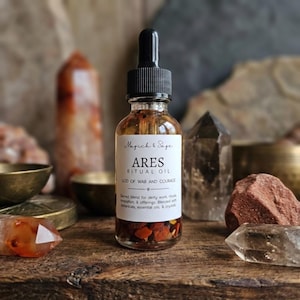 May include: A clear glass dropper bottle containing amber liquid and small red stones at the base. The white label reads Magick & Sage ARES RITUAL OIL GOD OF WAR AND COURAGE, with text describing a sacred blend for deity work, rituals, and offerings.
