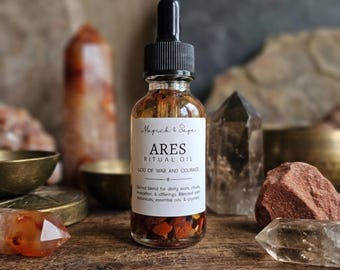 Op de afbeelding: Een doorzichtige glazen druppelfles met amberkleurige vloeistof en kleine rode steentjes op de bodem. Het witte etiket vermeldt Magick & Sage ARES RITUAL OIL GOD OF WAR AND COURAGE, met tekst over een heilige melange voor ritueel gebruik.