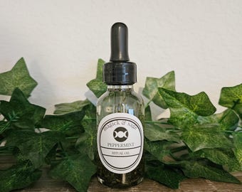 May include: A clear glass dropper bottle with a black cap containing liquid and herbs. The white label features the text "magick & sage", a triple moon symbol, "PEPPERMINT", and "RITUAL OIL" in black ink. The bottle is filled with a dark liquid.