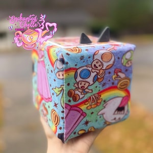May include: A blue and pink fabric cube with a colorful print of Mario characters, including a mushroom, a rainbow, and a star. The cube has a black felt top and a black felt bottom.