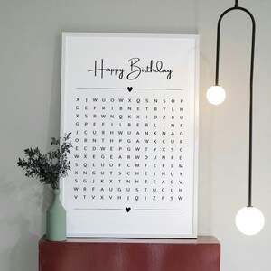 May include: A framed white print with the words "Happy Birthday" in cursive above a word search puzzle. The print is displayed on a red surface, with a green vase and black plant to the left. A modern light fixture hangs from the ceiling.