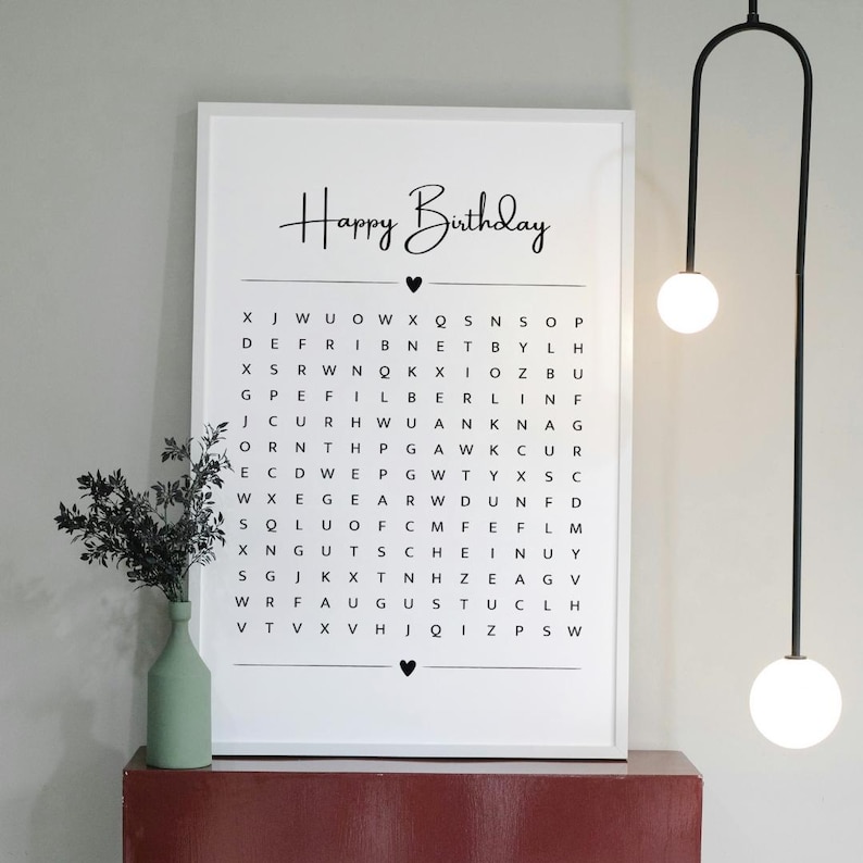 May include: A framed white print with the words "Happy Birthday" in cursive above a word search puzzle. The print is displayed on a red surface, with a green vase and black plant to the left. A modern light fixture hangs from the ceiling.