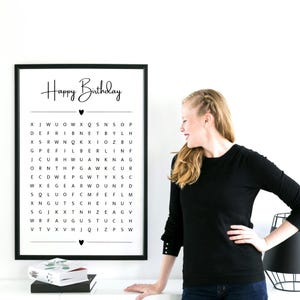 May include: A framed black and white word search puzzle with the words "Happy Birthday" at the top. The puzzle contains a grid of letters. A woman in a black long-sleeve shirt stands next to the framed puzzle.