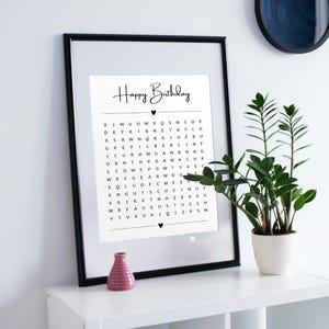 May include: A framed black and white print with the words "Happy Birthday" in cursive. Below the text is a word search puzzle. A small pink vase and a potted plant are on the white surface.