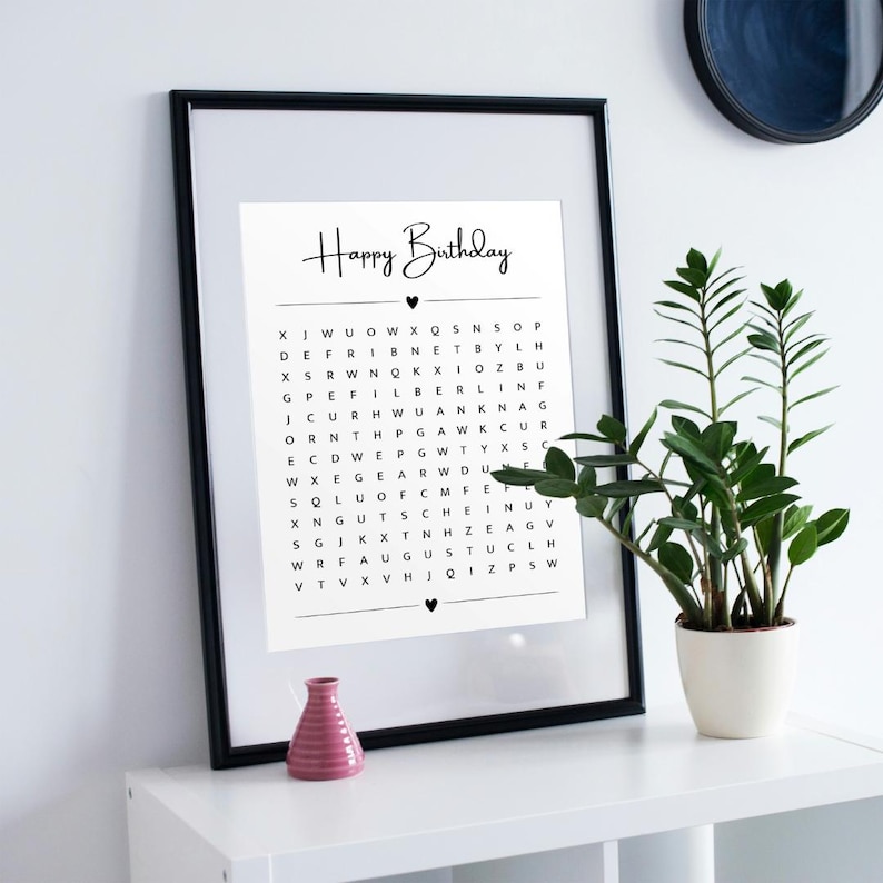 May include: A framed black and white print with the words "Happy Birthday" in cursive. Below the text is a word search puzzle. A small pink vase and a potted plant are on the white surface.
