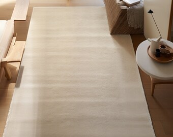 May include: A white, rectangular rug with a subtle texture. The rug is laid out on a wooden floor in a room with a large window and a wooden bench.