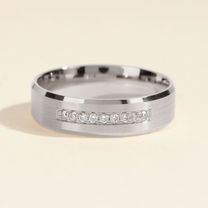 May include: A silver-toned metal ring featuring a brushed finish on the outer band and a central horizontal channel inlaid with a row of nine small, clear, round-cut stones. The edges of the ring are polished and beveled for a smooth look.