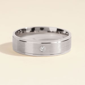 Op de afbeelding: Een zilverkleurige metalen ring met een geborstelde matte afwerking op de buitenste band en een enkele, kleine, ronde, heldere gefacetteerde edelsteen die verzonken in het midden is geplaatst. De randen van de band zijn gepolijst en reflecterend.