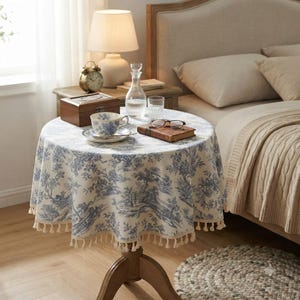 May include: A round table covered with a cream-coloured fabric featuring a blue toile de jouy pattern of trees and landscapes, finished with a fringe trim. The table holds a teacup, a saucer, a glass carafe, a book, and a wooden box.