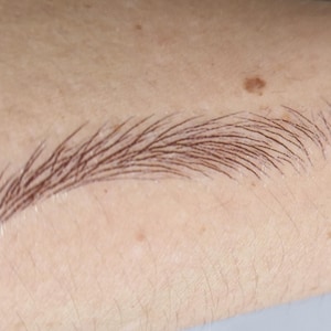 Può includere: Primo piano di un sopracciglio marrone disegnato sulla pelle. Il sopracciglio è arcuato e ha un aspetto naturale e sfumato. La pelle ha un tono chiaro con diversi piccoli nei.