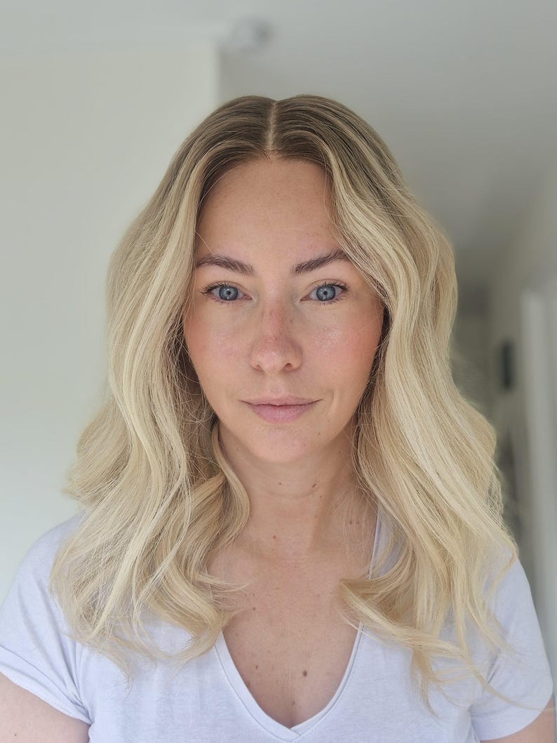 May include: Shoulder-length hair with a center part, featuring a bright blonde color with darker roots. The hair has loose, soft waves throughout the lengths and ends, creating a voluminous texture that frames the face in a natural, relaxed style.