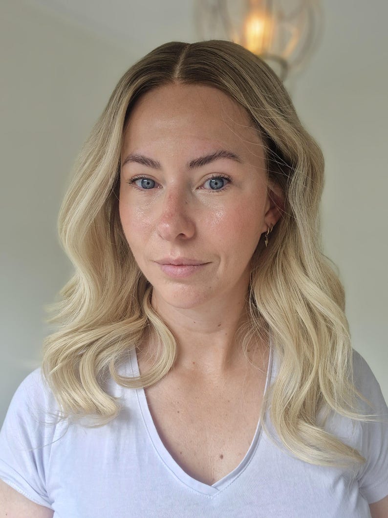 May include: A shoulder-length blonde wig with a center part and soft, loose waves. The hair features a natural-looking root transition from a darker shade to a bright, light blonde tone, styled with a smooth texture and a subtle, layered finish.
