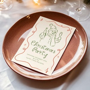 May include: A white invitation card with green and red text and a green line drawing of a snowman holding a bottle of champagne and a glass of champagne. The text reads "Christmas Party" and "Saturday, December 20th at 3 PM Silver Creek Inn Bend".