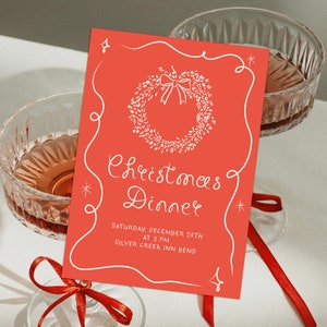 May include: A red invitation card with a white wreath and bow design. The text reads "Christmas Dinner" with the date and time "Saturday, December 20th at 3 PM" and the location "Silver Creek Inn Bend".