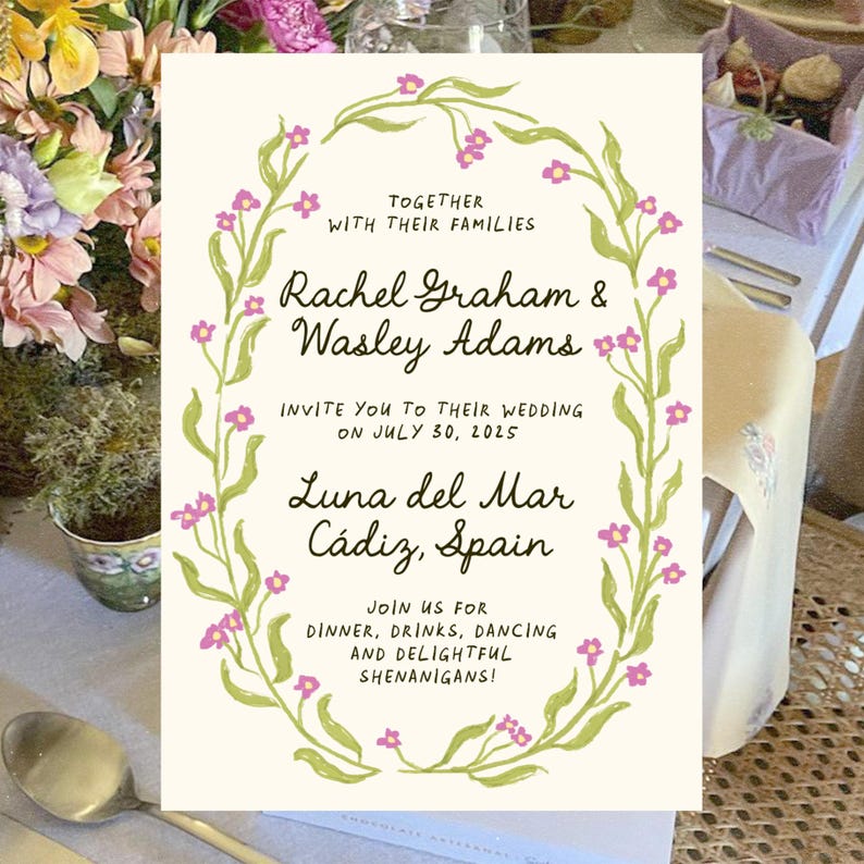 May include: A wedding invitation with a floral wreath border in shades of green and pink. The invitation reads "Together with their families, Rachel Graham & Wasley Adams invite you to their wedding on July 30, 2025, Luna del Mar, Cadiz, Spain. Join us for dinner, drinks, dancing and delightful shenanigans!"