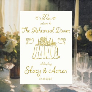May include: A white sign with gold lettering announcing "The Rehearsal Dinner" celebrating "Stacy & Aaron" on 08.10.2025. The sign features a hand-drawn illustration of a table setting with a floral centerpiece, candles, and two doves.