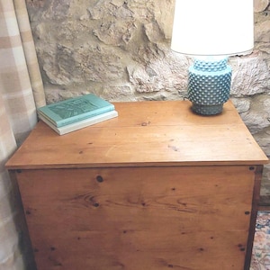May include: A wooden chest with a light brown finish, topped with books and a blue lamp with a white shade. The lamp has a textured base. The background is a stone wall.