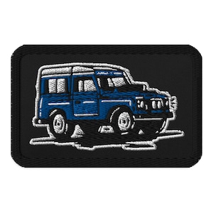 May include: Embroidered patch depicting a blue off-road vehicle with a white roof and trim. The patch has a black background and a white border, highlighting the vehicle's design. The patch is rectangular.
