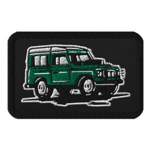 May include: An embroidered patch depicting a vintage Land Rover driving on a dirt road, in green and white.