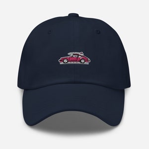 Puede incluir: Una gorra de béisbol azul marino con una imagen bordada en rosa de un coche con una tabla de surf en la parte superior.
