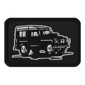 May include: A rectangular black embroidered patch featuring a white stitched outline of a boxy off-road vehicle. The design shows the side profile of the vehicle with detailed windows, wheels, and a front grille, all rendered in white thread on black.