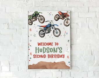 May include: A white birthday party sign with the text "Welcome to Hudson's Second Birthday" in green and red lettering. The sign is decorated with colorful stars and three cartoon dirt bikes.