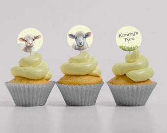 Puede incluir: Tres cupcakes con glaseado amarillo y adornos decorativos. Dos adornos presentan ilustraciones de acuarela de corderos sobre un fondo amarillo claro. El tercer adorno dice "Kimmy's Two" con un diseño de hierba. Los cupcakes están en moldes de papel grises.