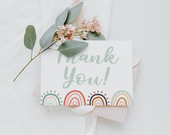 May include: A white card with the text "Thank You!" in green script. The card is decorated with a row of colorful rainbows at the bottom. The card is surrounded by pink flowers and green leaves.